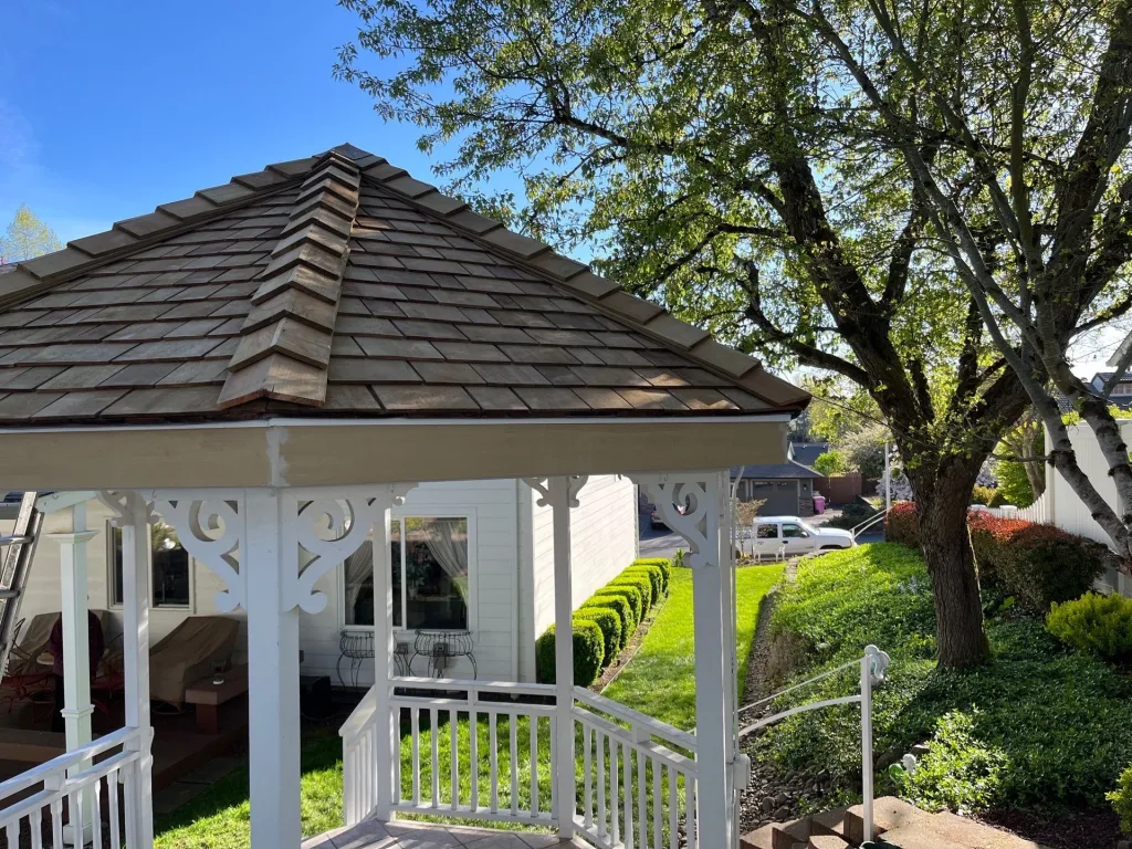 Replaced cedar shake gazebo roof with decorative ridge caps in a landscaped backyard with white railing and mature trees under a clear blue sky.